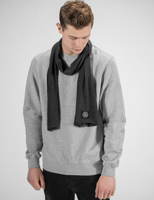 A man wearing a light gray crewneck sweatshirt and a dark gray knit scarf with a small circular logo patch, photographed against a light gray studio background.