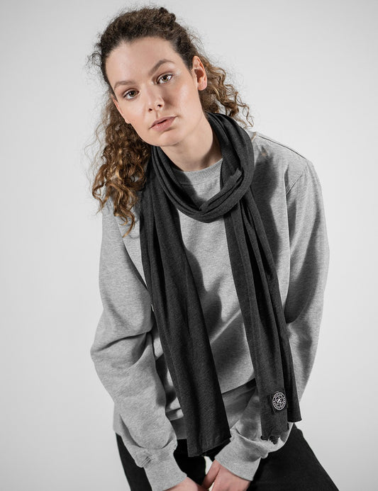 A woman with curly brown hair wearing a gray crewneck sweatshirt and a dark charcoal gray knit scarf with a small circular logo patch near the hem, posed against a light gray studio background.