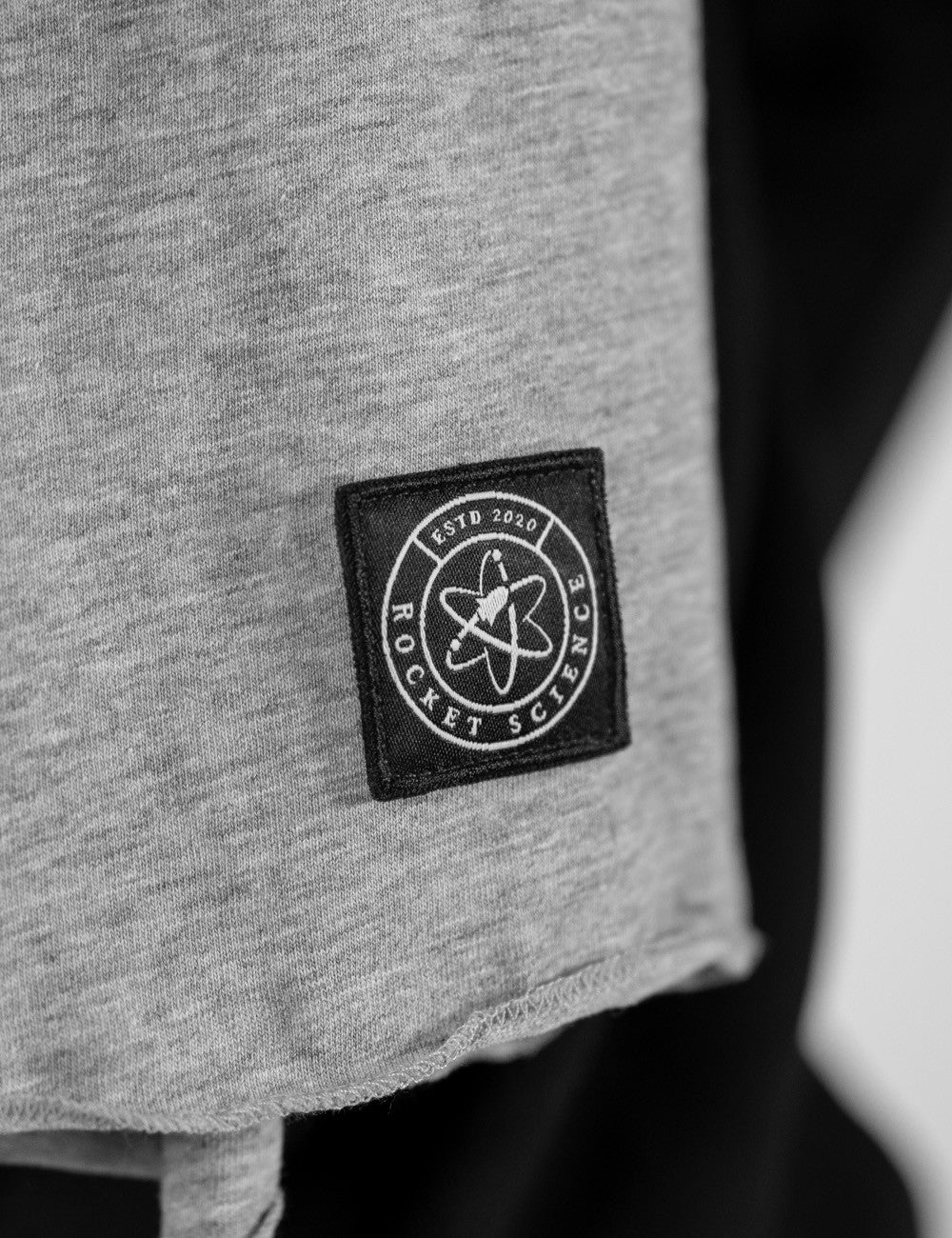 Close-up of the bottom corner of a light grey fabric scarf. A square black leather patch is sewn onto the fabric. The patch features a circular logo with an atom symbol in the center, a rocket ship icon, and the words "ESTD 2020" and "ROCKET SCIENCE" printed in white around the circle. The bottom edge of the grey scarf is frayed and slightly rolled. 