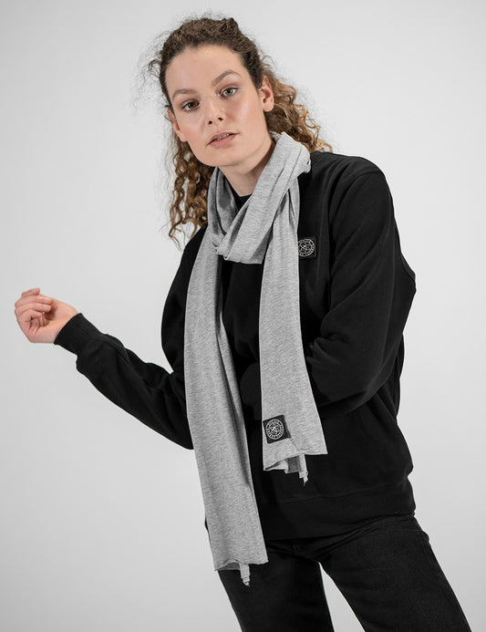 A young woman wearing a light grey jersey fabric scarf draped around his neck over a solid black crewneck sweatshirt and dark jeans. The scarf has a small, square dark Rocket Science logo patch near the bottom edge.