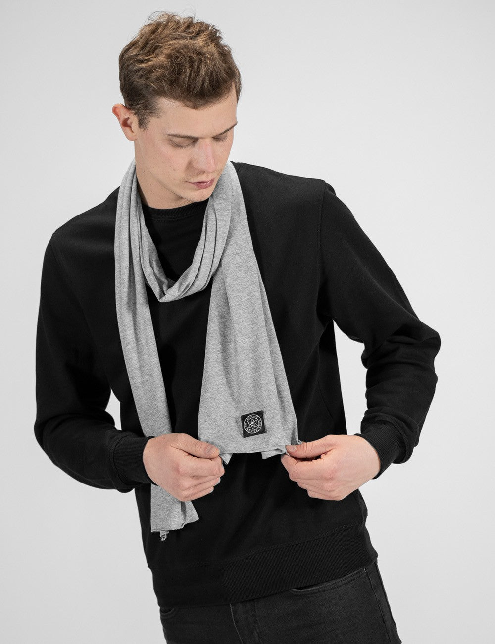 A young man looking downward, wearing a light grey jersey fabric scarf draped around his neck over a solid black crewneck sweatshirt and dark jeans. He is holding both ends of the scarf with his hands near his chest. The scarf has a small, square dark "Rocket Science" logo patch near the bottom edge. 