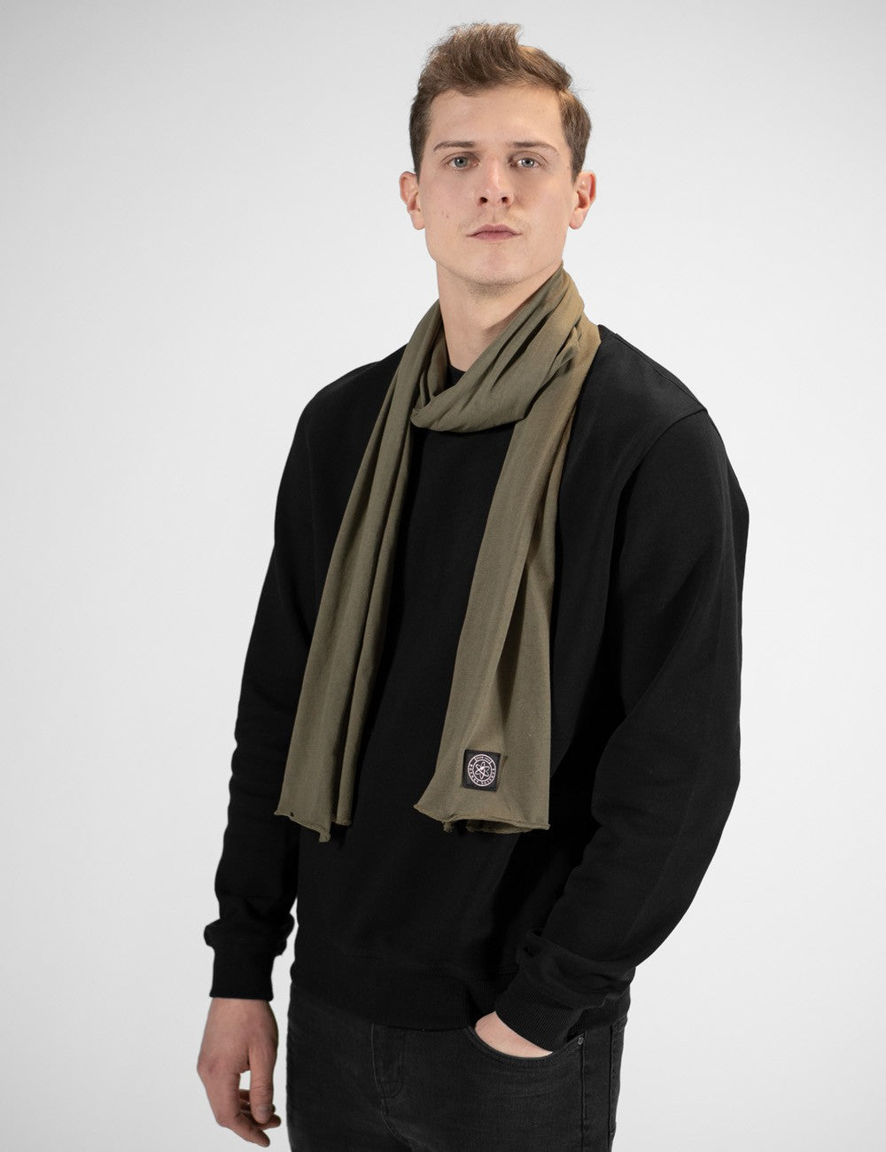 A close-up view highlighting an olive green jersey scarf from Rocket Science, loosely wrapped around a man's neck. The fabric hangs smoothly down the front of a black sweatshirt. A prominent detail is the small, square dark logo patch stitched near the bottom edge of the scarf's right end.
