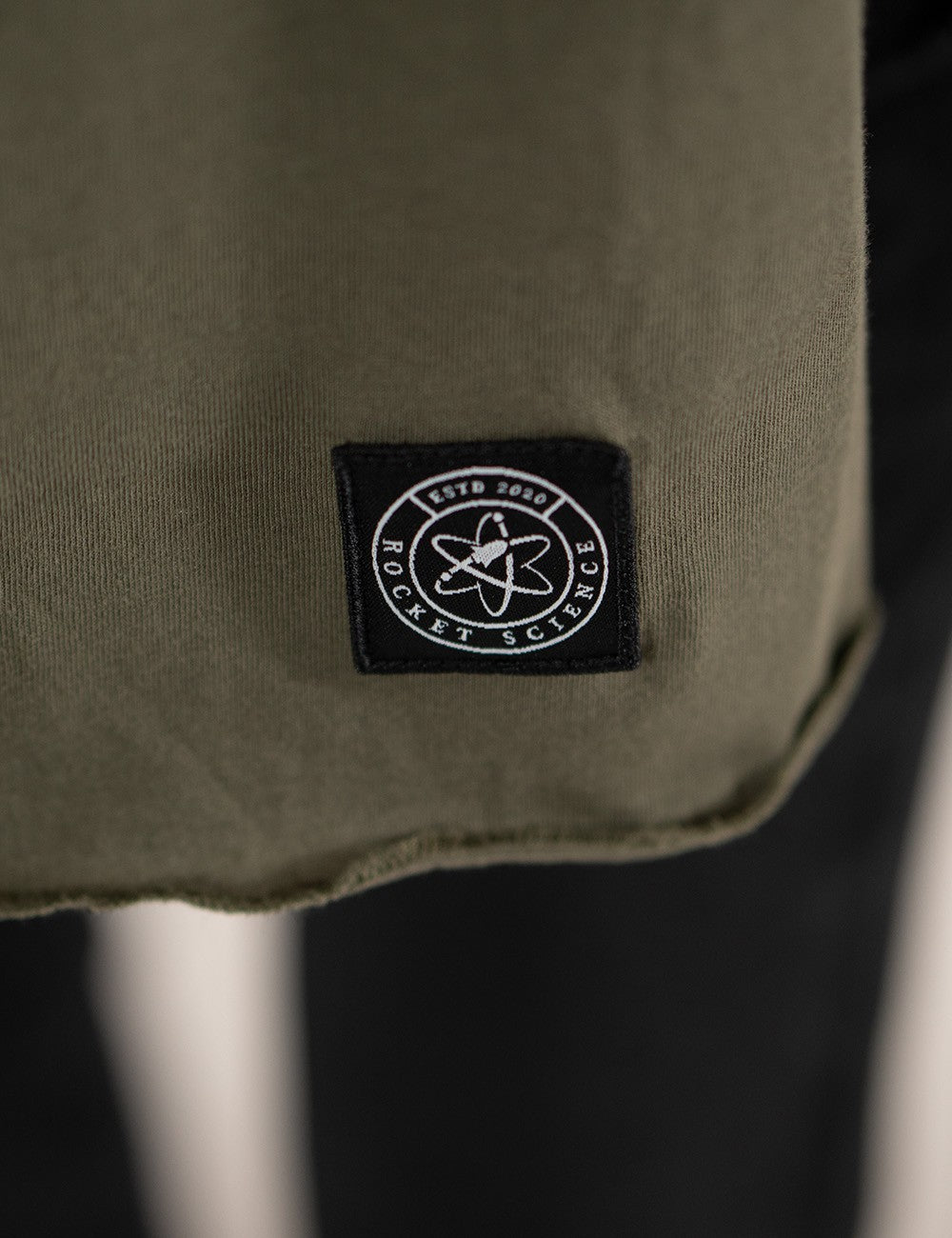 A close-up view highlighting an olive green jersey scarf from Rocket Science. A prominent detail is the small, square dark logo patch stitched near the bottom edge of the scarf's right end.