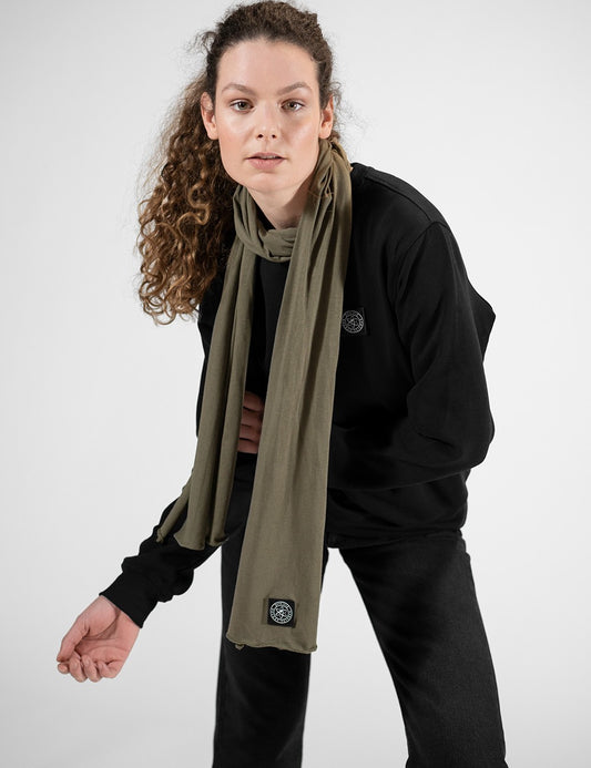 A close-up view highlighting an olive green jersey scarf from Rocket Science, loosely wrapped around a woman's neck. The fabric hangs smoothly down the front of a black sweatshirt. A prominent detail is the small, square dark logo patch stitched near the bottom edge of the scarf's right end.