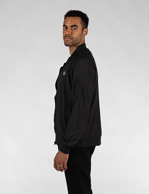 A man in a black jacket and black pants, shown in profile against a light gray studio background.