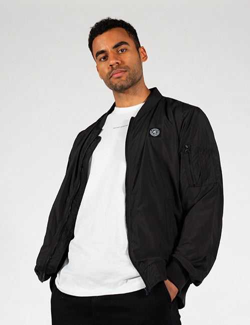 A man wearing a black bomber jacket over a white t-shirt and black pants, standing against a light gray studio background.
