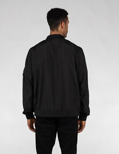 A man viewed from behind wearing a black bomber jacket and black pants against a light gray studio background.