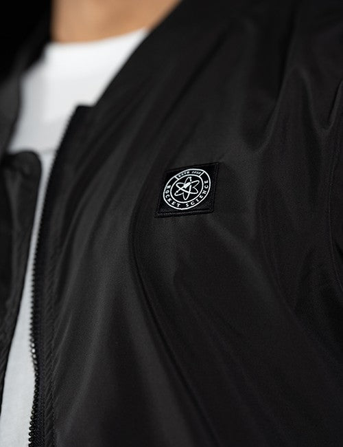 Close-up of a black zip-front jacket worn over a white shirt, featuring a circular black-and-white Rocket Science logo patch on the chest.