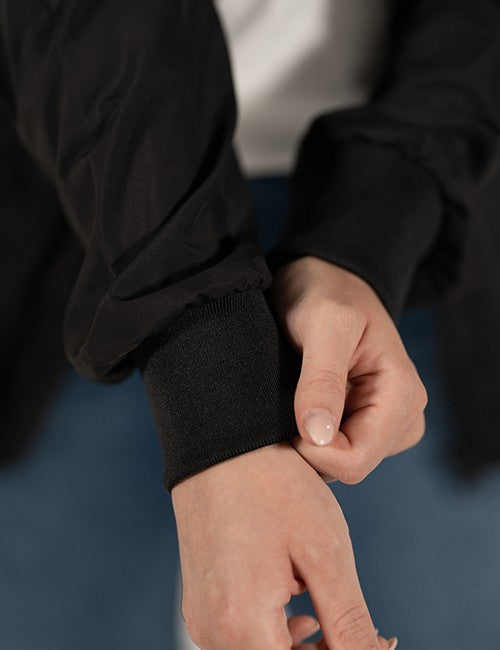 Close-up of a person's hands adjusting the ribbed knit cuff of a black long-sleeve jacket, with a white shirt and blue jeans visible in the background.