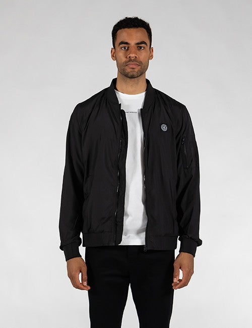 A man wearing a black bomber jacket over a white t-shirt and black pants, standing against a light gray studio background.