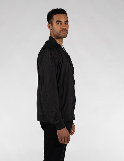 A man wearing a black long-sleeve zip-up jacket and black pants, shown in profile against a light gray studio background.