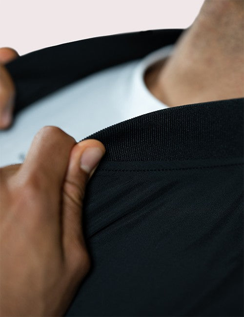 Close-up of a person pulling a black ribbed waistband or hem away from their body, revealing a white undershirt. The hand and lower torso are visible, with a plain light background.