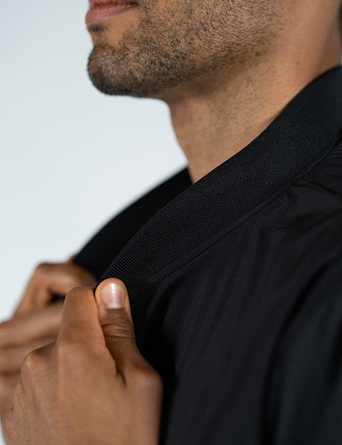 Close-up of a man wearing a black collared shirt, adjusting the collar with his hand against a light gray background.