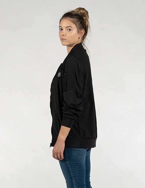 A woman wearing a black bomber jacket and blue jeans, shown from a side profile against a light gray studio background.