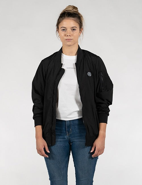 A woman modeling a black bomber jacket over a white t-shirt and blue jeans, standing against a light gray studio background.