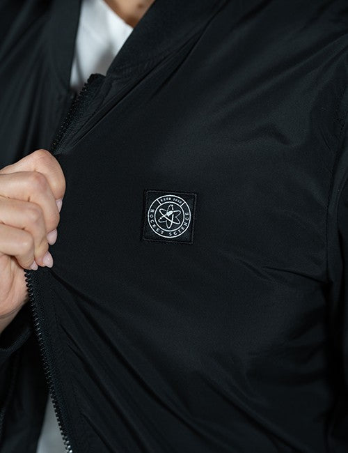 Close-up of a black zip jacket being opened by a hand, featuring a circular black-and-white Rocket Science logo patch on the chest.