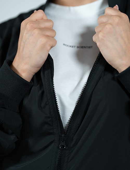 A close-up of a person wearing a black zip-up jacket over a white t-shirt with the text 'ROCKET SCIENTIST' printed on the chest, with both hands pulling the jacket open.