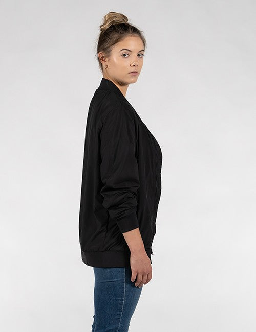 A woman shown in side profile wearing a black bomber jacket and blue jeans against a plain light gray studio background.