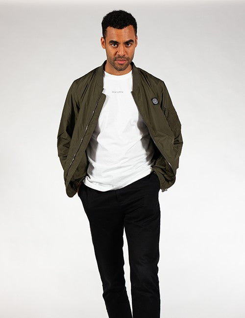 A man standing against a plain gray background wearing an open olive green bomber jacket over a plain white t-shirt and black pants.