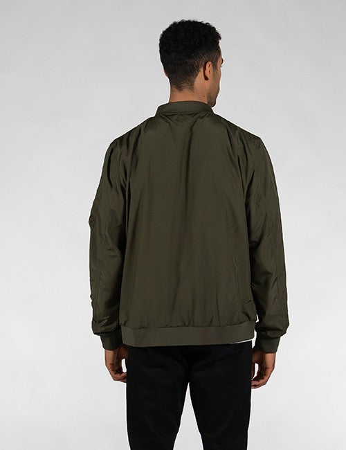 A man viewed from the back wearing an olive green bomber jacket and black pants, standing against a light gray studio background.