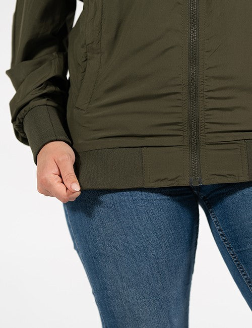 Close-up of a person wearing an olive green zip-front jacket with blue jeans against a plain light background.