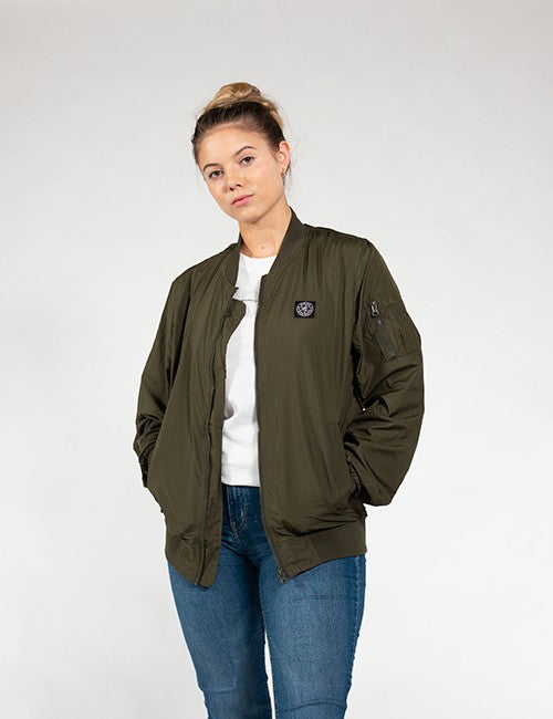 A woman modeling an olive green bomber jacket over a white t-shirt and blue jeans, standing against a light gray studio background.