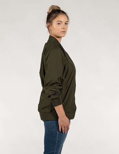 A side profile view of a woman with her hair in a high bun, wearing a dark olive green satin-finish bomber jacket and blue jeans against a plain off-white background.