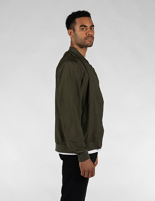 A man shown in profile wearing an olive green bomber jacket and black pants against a light gray studio background.