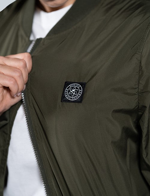 Close-up of a person wearing an olive green bomber jacket, pulling the zipper open with a hand. A black square patch with a white Rocket Science logo is visible on the chest, and a white shirt is underneath.