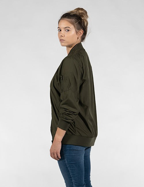 A side profile view of a woman with her hair in a high bun, wearing a dark olive green satin-finish bomber jacket and blue jeans against a plain off-white background.