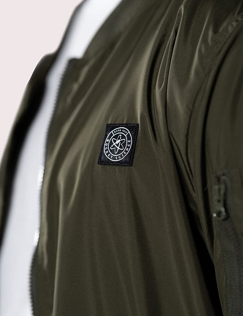 A close-up shot of an olive-green bomber jacket showing its glossy, water-resistant fabric. A black square patch is stitched onto the chest, featuring a white circular Rocket Science logo with an atomic orbital design in the center. A vertical zipper is visible on the left side of the frame, and a small zippered utility pocket can be seen on the sleeve to the right.