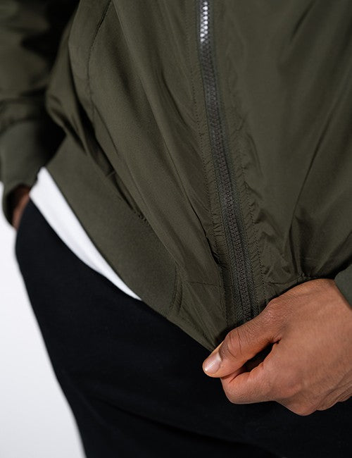 Close-up of a person wearing an olive green zip-front jacket over a white shirt and black pants, with the hand pulling the jacket open.