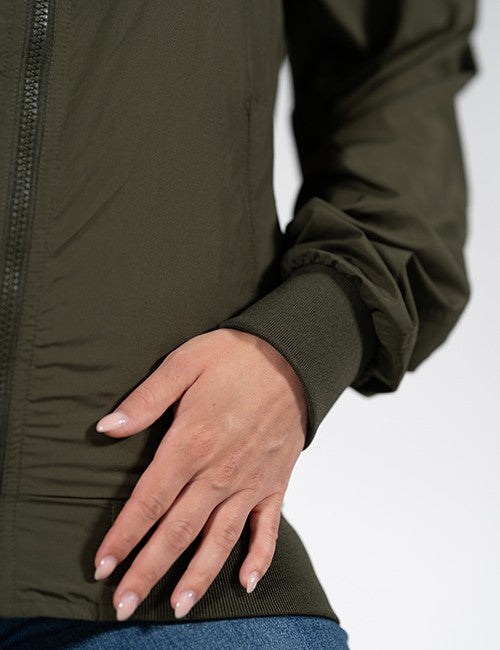 Close-up of a person wearing an olive green zip-front jacket with blue jeans against a plain light background.