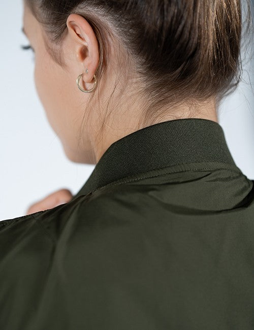 An over-the-shoulder, close-up shot from behind shows a person with brown hair tied up in a high bun. They are wearing a dark olive green bomber jacket with a ribbed collar.