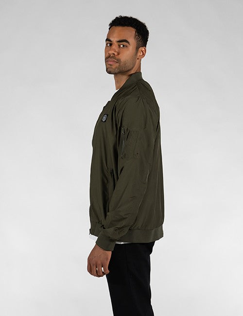 A man wearing an olive green bomber jacket, shown in profile against a light gray studio background.