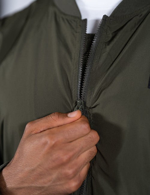 A close-up shot of a person's hand pulling up the dark zipper of an olive green bomber jacket.