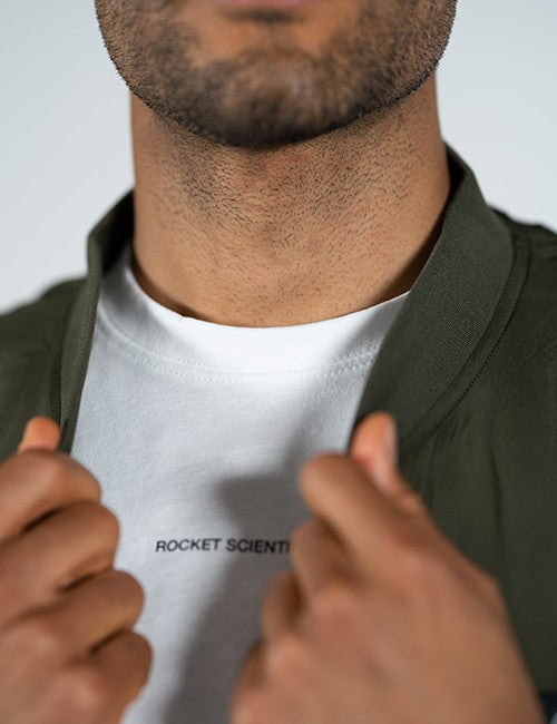 Close-up of a man wearing a white crewneck t-shirt with 'ROCKET SCIENTIST' printed in black on the chest, layered under an olive green jacket.