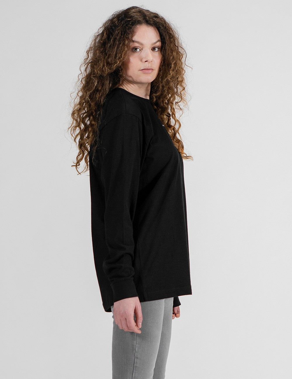 A woman with long, voluminous, curly brown hair stands in a side-profile view against a solid grey background. She is wearing a loose-fitting, solid black long-sleeve crewneck shirt and grey pants.