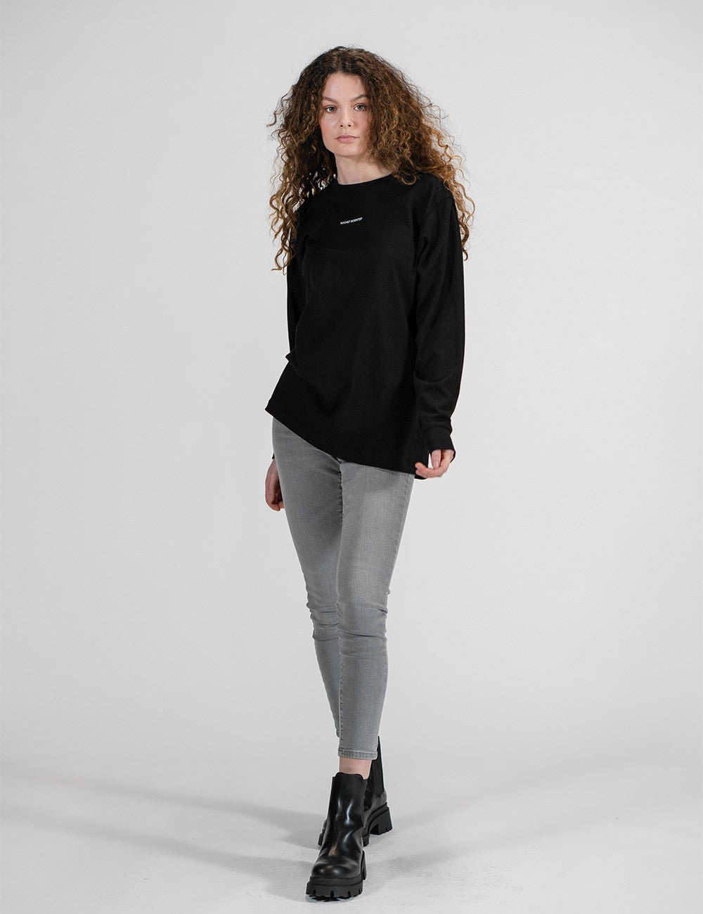 This image shows a young woman wearing a black long-sleeve shirt with a small "Rocket Scientist" chest print , paired with gray skinny jeans and chunky black boots.