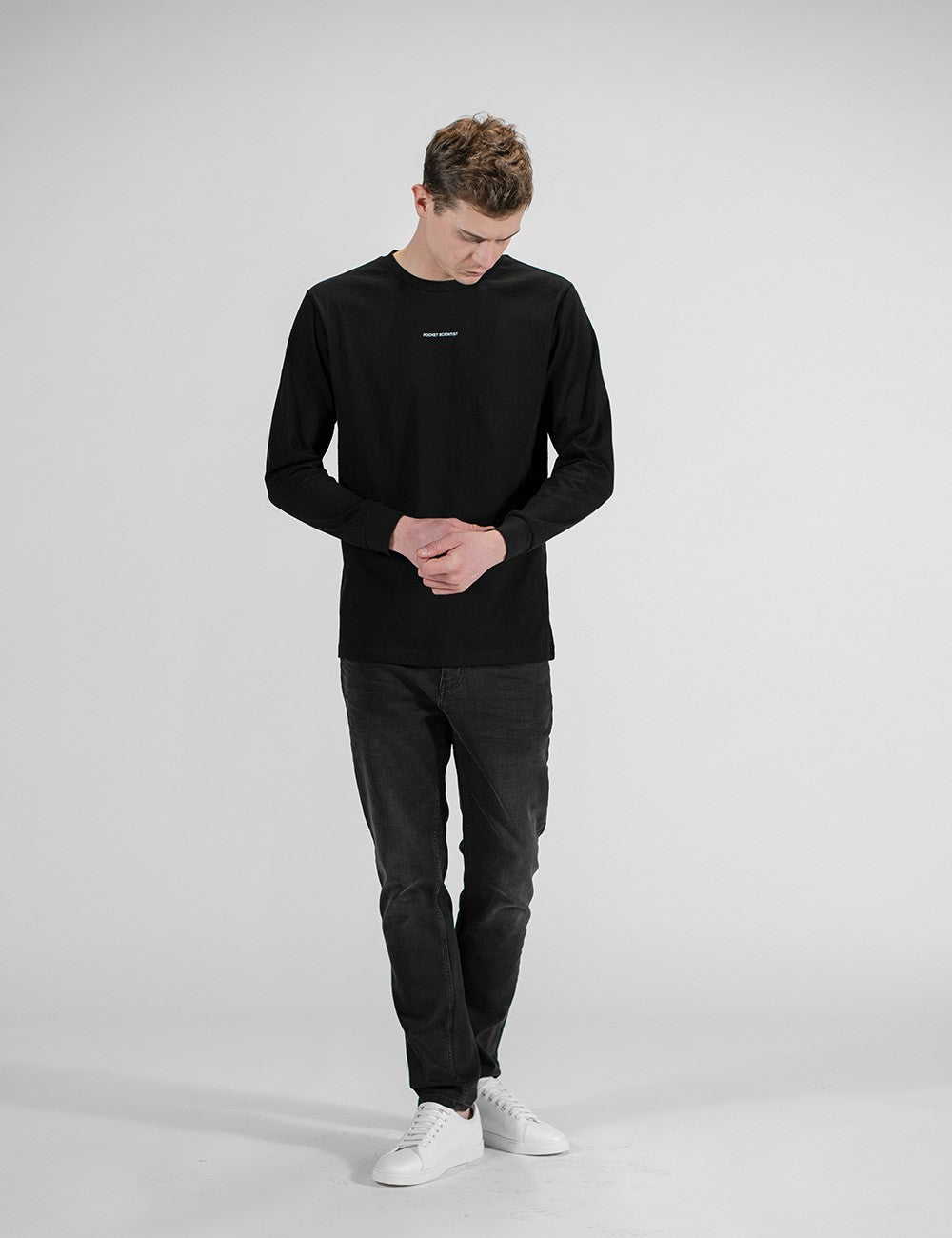 This image shows a front pose of a young man wearing a plain black long-sleeve crewneck shirt with a small logo Rocket Scientist print on the chest , with dark denim jeans and white sneakers.