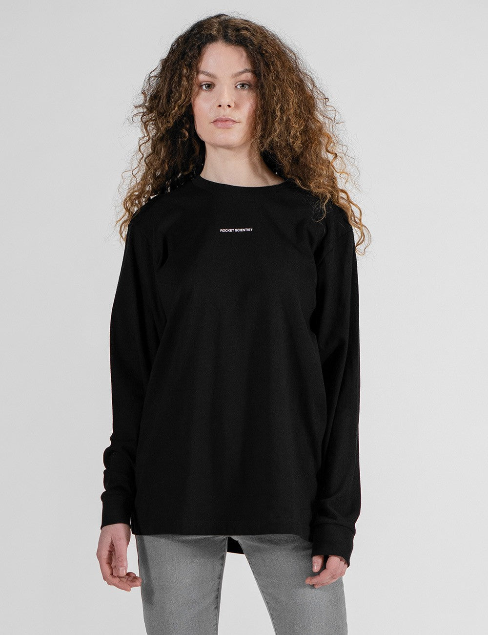 This image shows a front pose of a young woman wearing a plain black long-sleeve crewneck shirt with a small logo Rocket Scientist print on the chest 