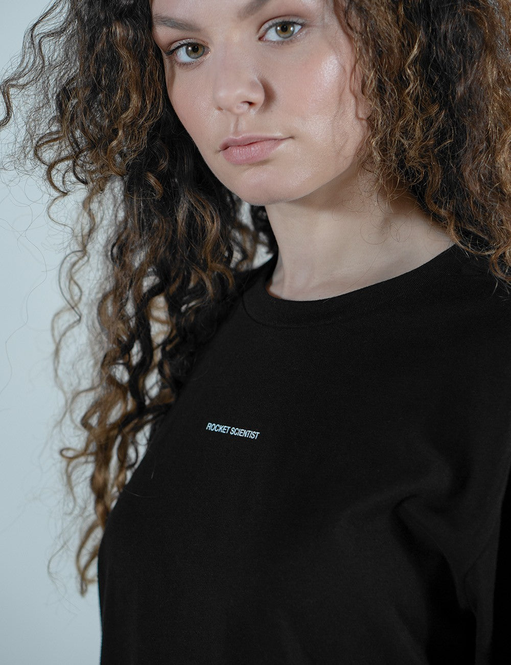 A close-up shot of a young woman with long, curly brown hair looking at the camera. She is wearing a plain black crewneck shirt with the words "ROCKET SCIENTIST" printed in small, white, all-caps letters across the center of the chest.