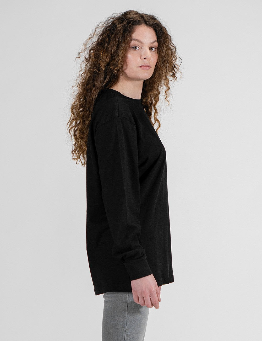 A model with long, curly brown hair is photographed from a side profile against a plain white background. She is wearing an oversized, black long-sleeve crewneck shirt and grey pants.