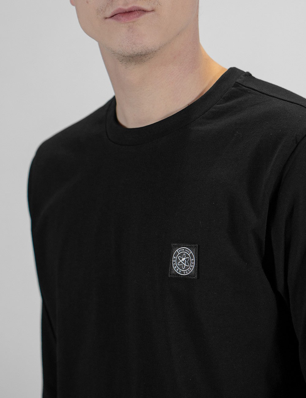 A close-up shot of a man wearing a black round-neck long-sleeve sweatshirt. The garment features a small, square, black-and-white circular graphic patch stitched onto the left side of the chest. The logo displays an atomic or planetary orbit ring symbol inside a circle with text wrapping around it. The background is a plain, neutral light gray.