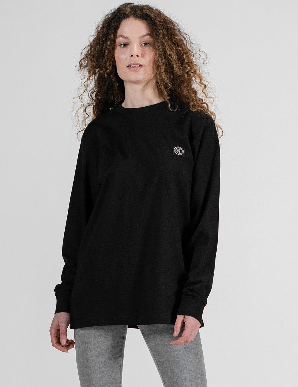 A woman with long, curly brown hair is photographed from a front profile against a plain white background. She is wearing an oversized, black long-sleeve crewneck shirt with Rocket Science logo patch and grey pants.