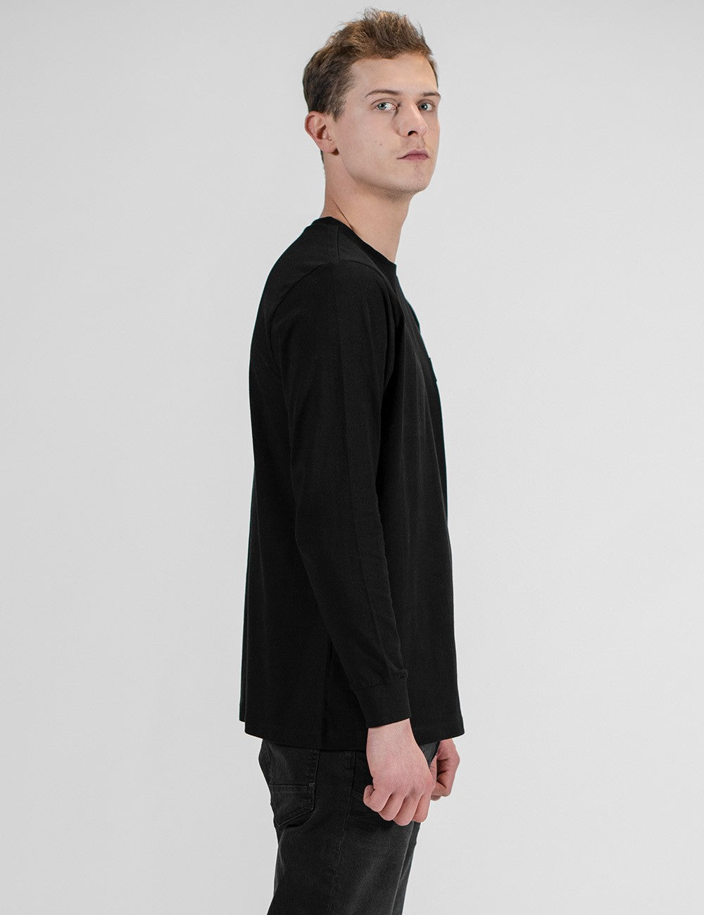 A model with light brown hair standing in profile, wearing a black long-sleeved t-shirt and black pants against a plain grey background.