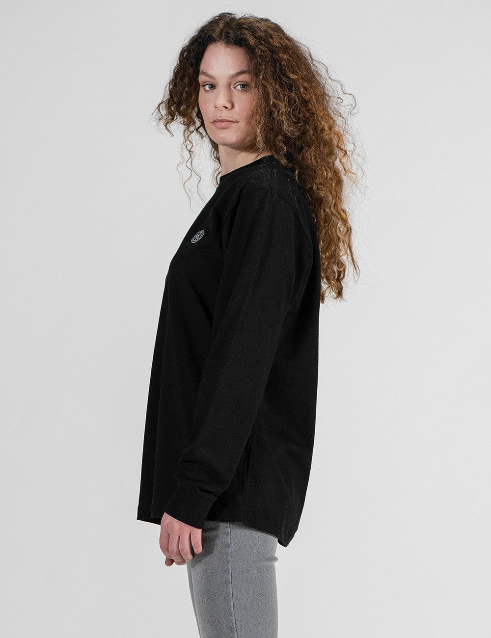 A woman with long, curly brown hair is photographed from a side profile against a plain white background. She is wearing an oversized, black long-sleeve crewneck shirt and grey pants.