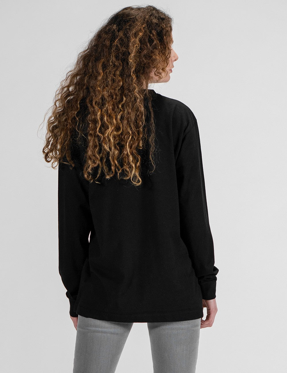 A woman with long, curly brown hair is photographed from a rear pose against a plain white background. She is wearing an oversized, black long-sleeve crewneck shirt and grey pants.