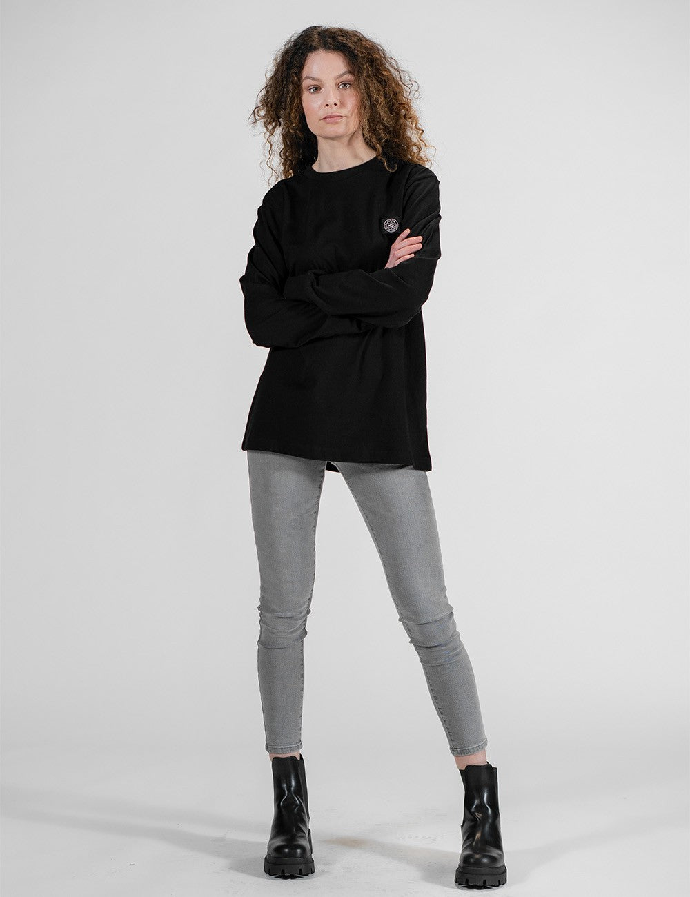 A full-length studio portrait of a woman with long, curly brown hair standing with her arms crossed against a plain grey background, wearing a loose-fitting black long-sleeve shirt with a small circular chest logo, grey skinny jeans with knee seams, and chunky black leather platform ankle boots.