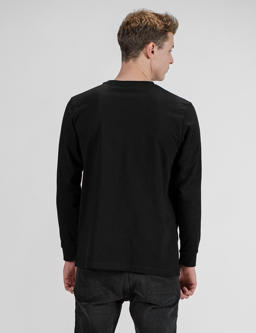 A young man with light brown, wavy hair is shown from the back, wearing a plain black long-sleeved crewneck shirt and black jeans.